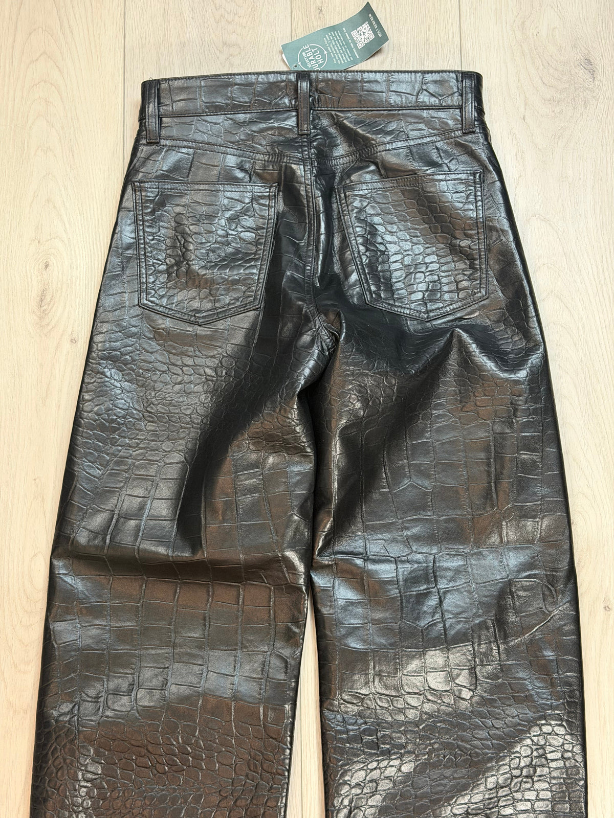 Black crocodile-embossed leather pants laid flat on a light wood floor, showing the front pockets and waistband details.